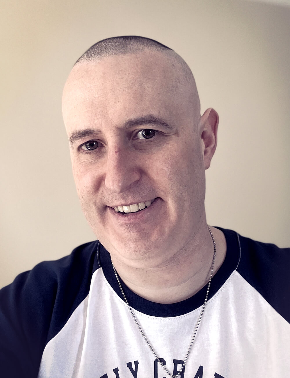 A person wearing a black and white shirt and a necklace, smiling at the camera with a close-cropped haircut.